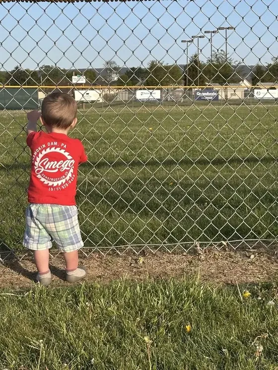 A toddler in a red Smego Construction Co. t-shirt at the fence of a ball field in Shamokin Dam, Pennsylvania