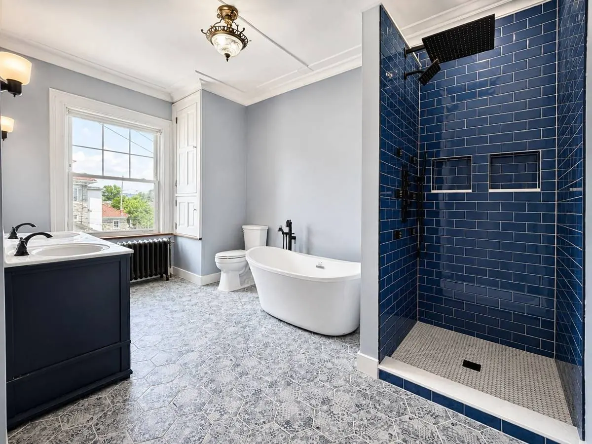 Restored primary bathroom in a historic Sunbury, Pennsylvania home, with a freestanding tub, cobalt blue tiled shower, patterned hexagon floor and the original cast iron radiator