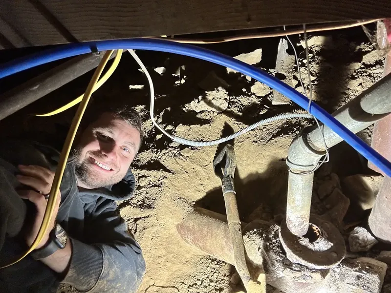 Mike Smego in a crawlspace beneath a house, new PEX water lines run alongside the original cast iron drain